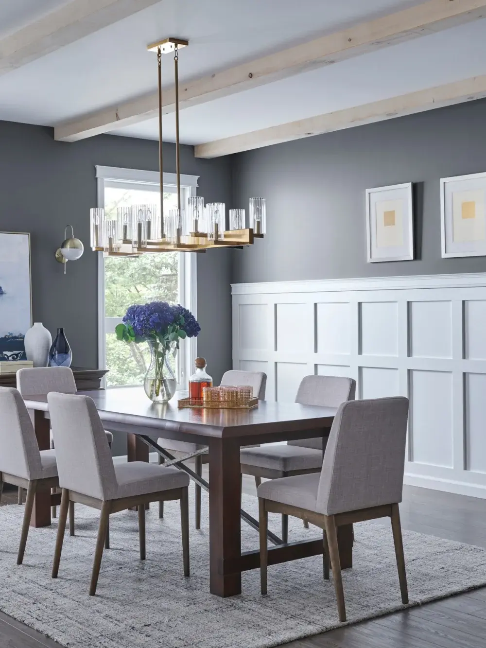 An elegant dining room with medium gray walls and white board and batten styled wainscot. In the center of the room is a simple wood dining table surrounded by chairs with a large area rug underneath.