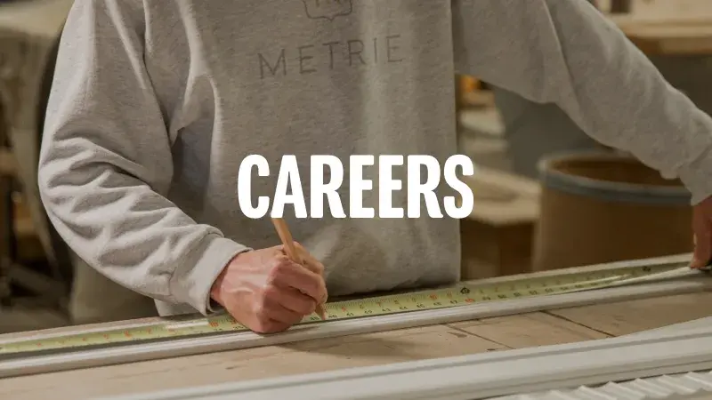 A man in a grey 'Metrie' sweatshirt measures a piece of moulding in a workshop. The text "Careers" is overlaid over top the image.
