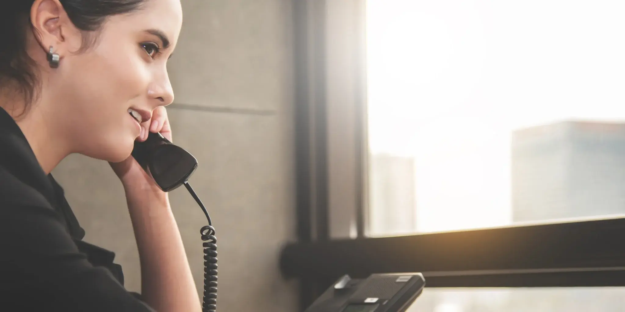 A young Caucasian woman in a black dress shirt is talking on a black corded telephone.